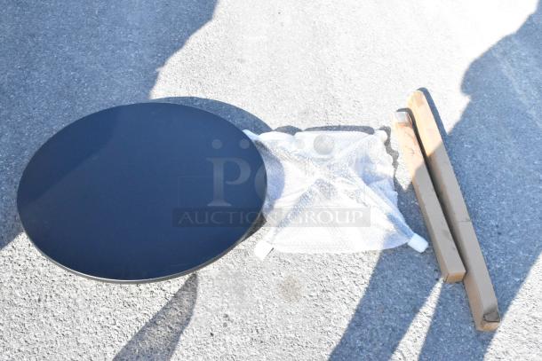 Round black table top with two packaged leg components and bubble-wrapped base on concrete. Assembly required.