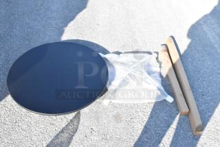 Round black table top with two packaged leg components and bubble-wrapped base on concrete. Assembly required.