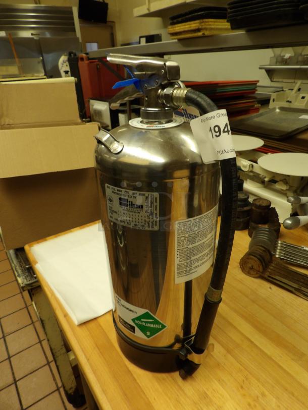 Stainless steel fire extinguisher on a wooden table, featuring inspection labels and a non-flammable gas tag.