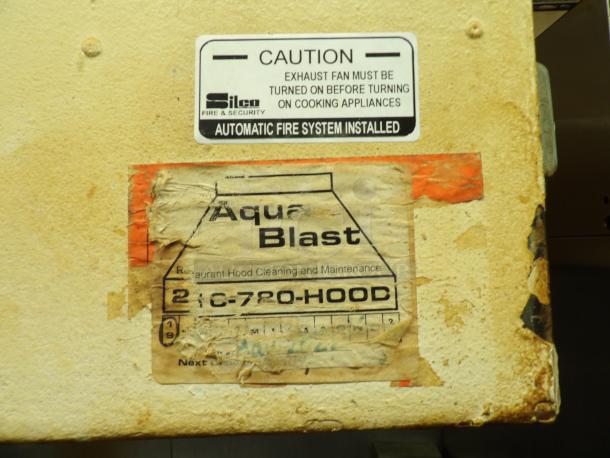 Worn "Aqua Blast" label on restaurant hood cleaner with caution for exhaust fan use; automatic fire system installed.