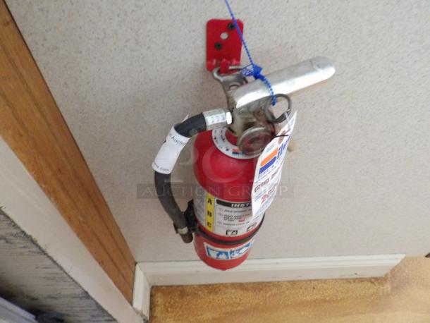 Red fire extinguisher wall-mounted, with attached inspection tags. Labels display usage instructions and safety icons.