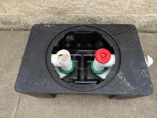 Black plastic tank with circular opening, two white PVC pipes, and one red cap. Surface shows wear.