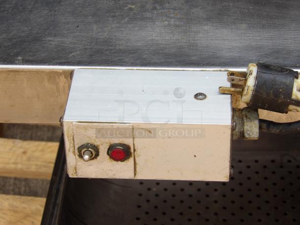Metal electrical box with toggle switch and red indicator light, attached to a stainless steel surface. Visible power plug connection.
