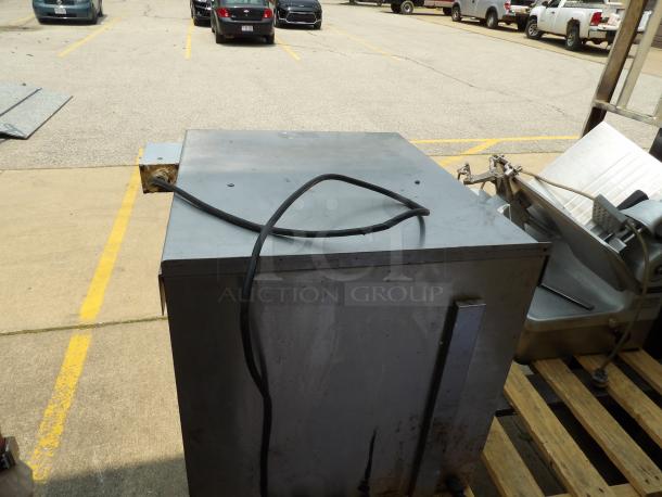 Commercial stainless steel meat slicer on a wooden pallet, showing signs of wear. Power cord visible; no labels or markings.
