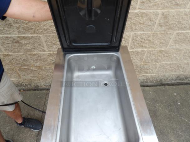 Stainless steel ice cooler, open lid showing large compartment and drainage hole, in good condition, with outdoor setting.