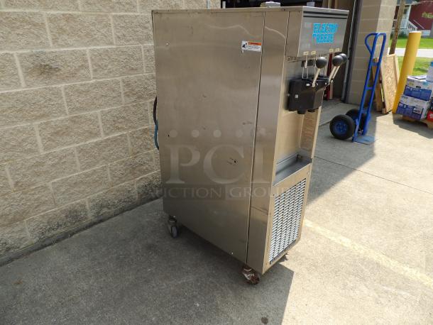 Commercial Electro Freeze soft serve machine, stainless steel, dual levers, on wheels, scuffs on surface, outdoor setting.