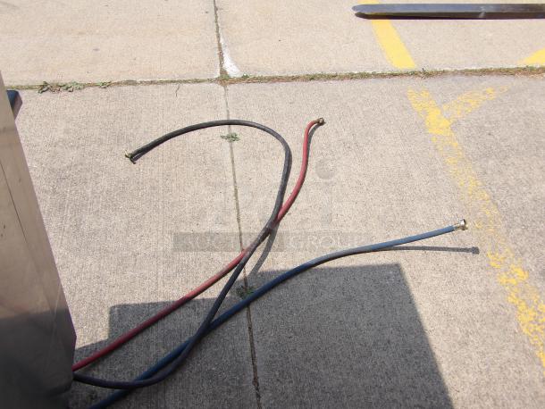Two rubber hoses in red and black on pavement, with metal connectors. Used condition, suitable for various applications.