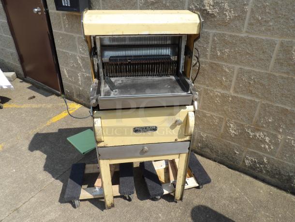 Vintage Oliver bread slicer, cream-colored, showing wear. Metal components, safety slicer label visible. Urban industrial setting.