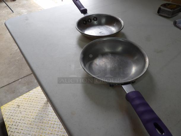 Two well-used frying pans with purple handles, showing minor scratches. No visible labels or markings.
