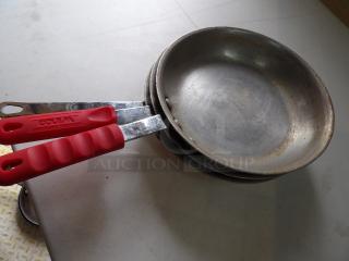 Two metal pans with red rubber handles, marked "Winco," showing light use and wear on a white surface.