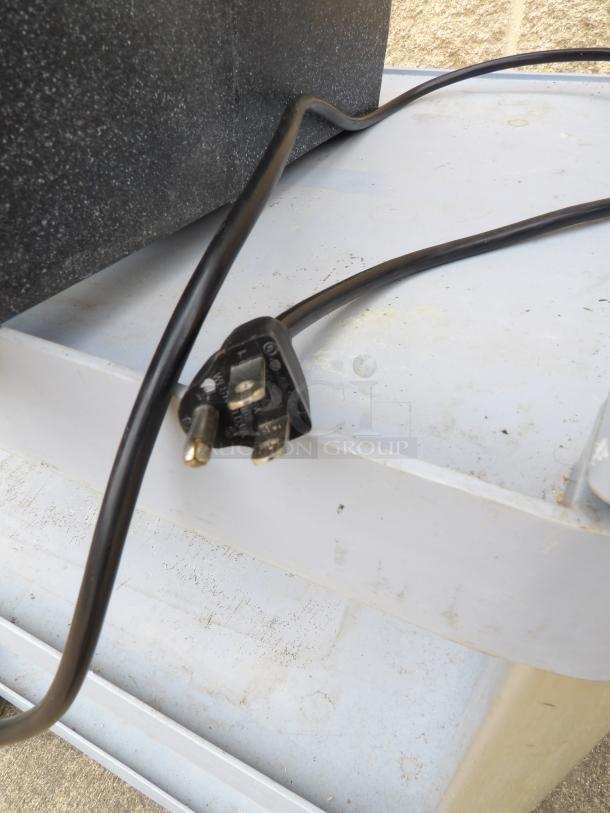 Black power plug with three prongs and long cord, attached to a black device on a metal shelf. Condition appears used.