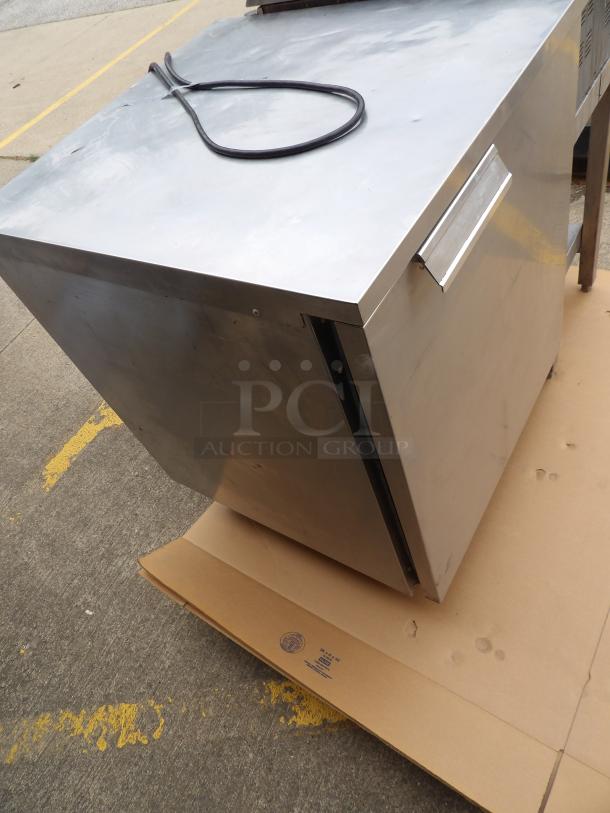 Stainless steel commercial refrigerator with handle, good condition, resting on cardboard. Power cord visible.