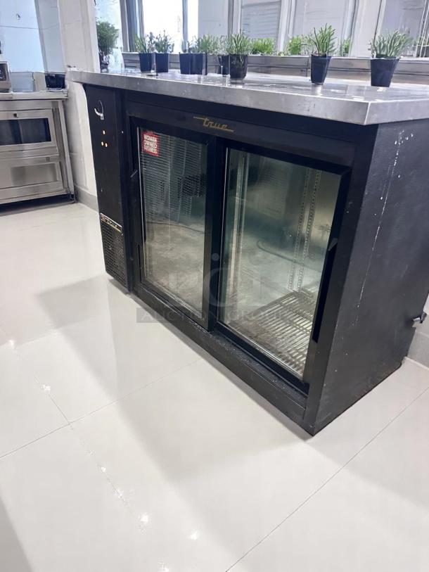 True refrigerated display cooler, dual glass doors, black, stainless steel top, good condition, "Woman Owned Business" sticker.