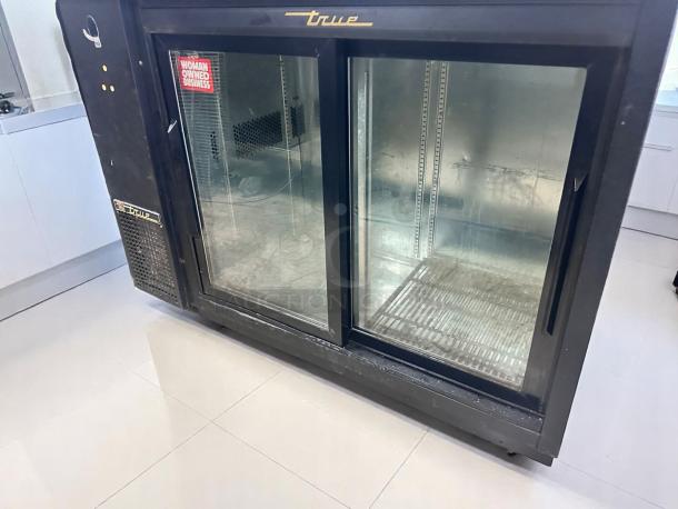 True commercial display refrigerator, black with sliding glass doors. Features a "Woman Owned Business" sticker. Signs of wear.