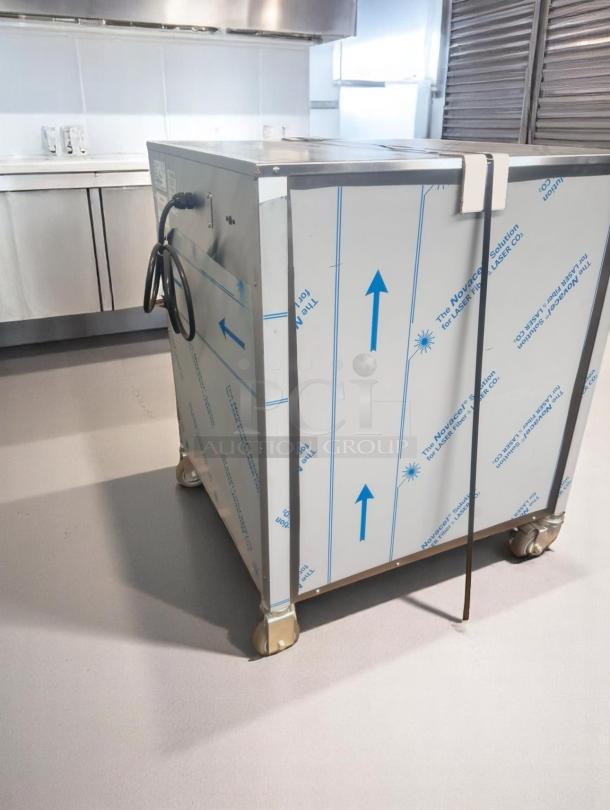 Commercial stainless steel kitchen appliance, protective film with arrows and branding. New condition on caster wheels.