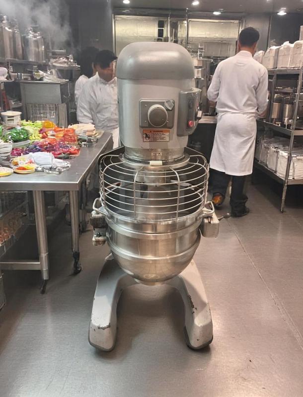 Commercial kitchen stand mixer, large capacity, stainless steel bowl, visible wear on base, safety guard feature.