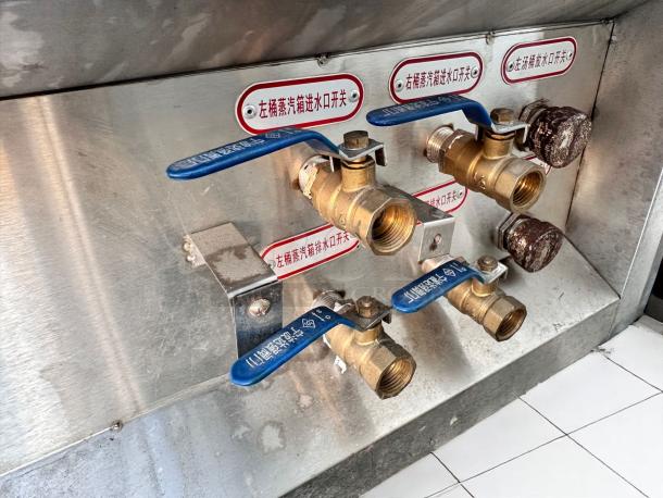 Industrial valve assembly with three brass ball valves, blue levers, and labeled inlets on a stainless steel panel. Good condition.