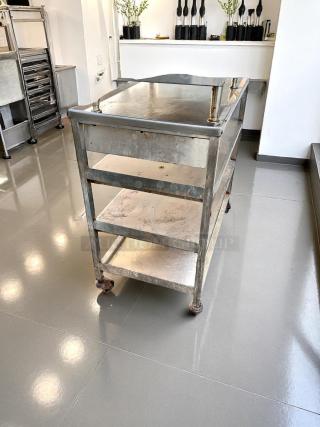 Stainless steel utility cart with three shelves, signs of wear, and casters for mobility. Ideal for kitchen or industrial use.