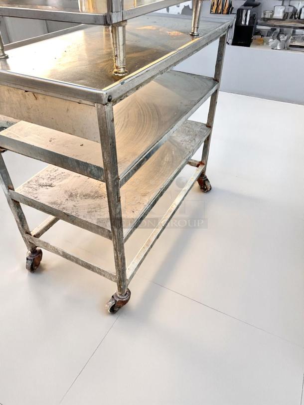 Stainless steel three-tier industrial cart with wheels. Shows wear and rust, ideal for kitchen or restaurant use.