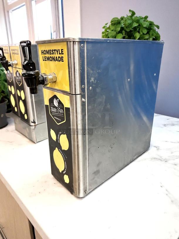 Stainless steel lemonade dispenser with "Homestyle Lemonade" label. Features black spigot, minor scuffs, plant on top.