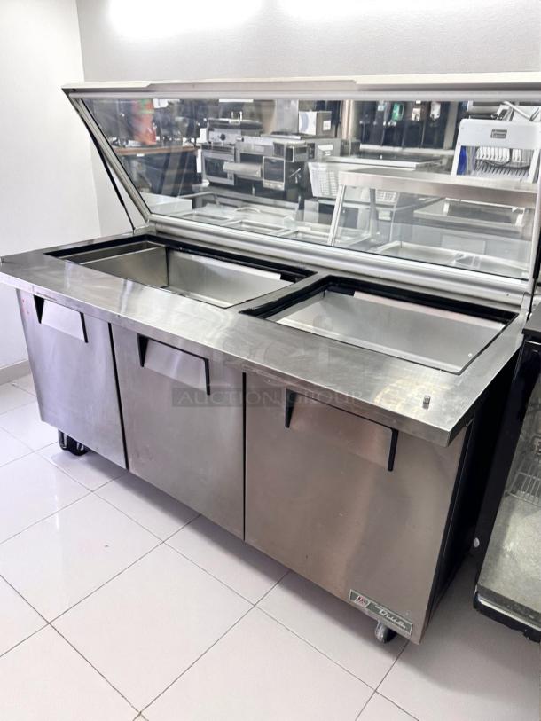 Commercial stainless steel refrigerated prep table with glass top, three compartments, clean condition, wheels for mobility.