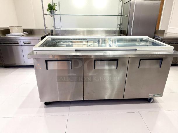 Three-door stainless steel refrigerated display counter on wheels, glass top, True brand, good condition, for commercial use.