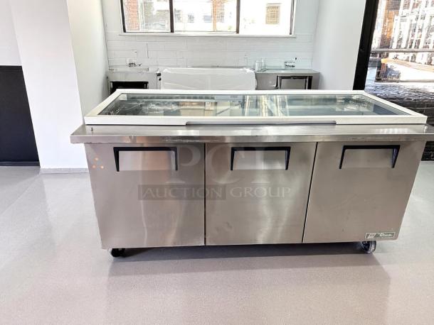 Stainless steel commercial refrigeration unit with three compartments, glass top, and caster wheels. Brand: True, in good condition.