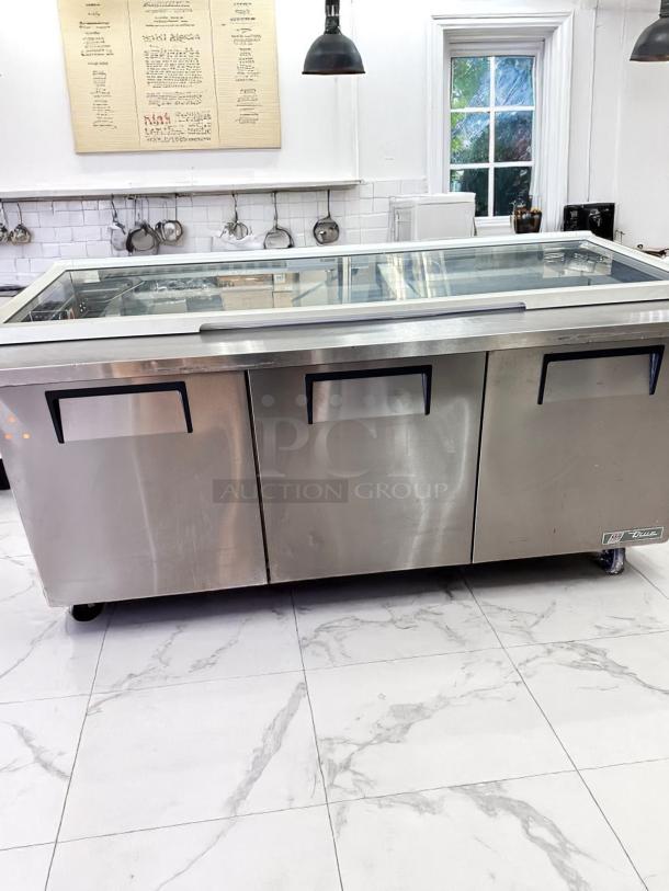 Commercial stainless steel refrigeration unit with a glass top, three doors, and visible brand logo "True." Used condition.