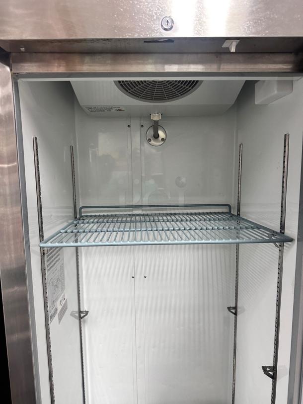 Commercial stainless steel refrigerator interior, single wire shelf, adjustable rack system, fan and light fixture visible.