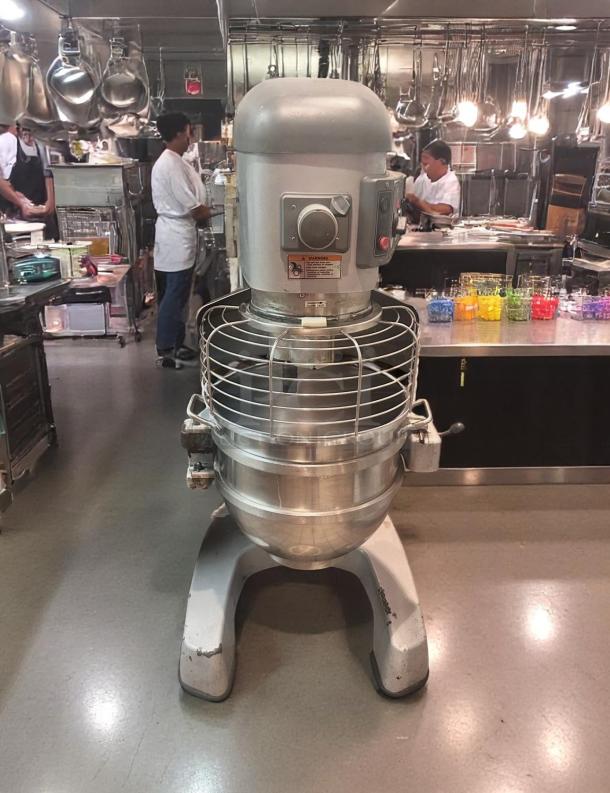 Industrial stand mixer with large stainless steel bowl, safety cage, control panel, and warning label. Used condition.