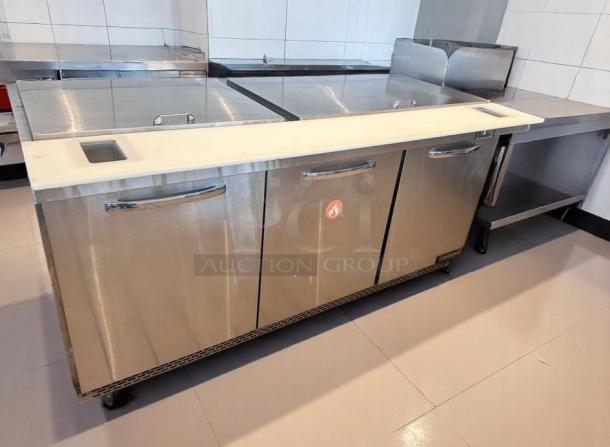 Stainless steel commercial kitchen refrigeration unit with three doors, cutting board top, and flame logo. Excellent condition.