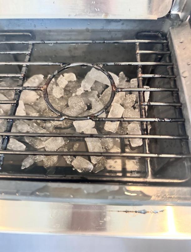 Stainless steel ice maker filled with clear ice cubes, showing metal grid pattern inside. In good condition with visible water droplets.