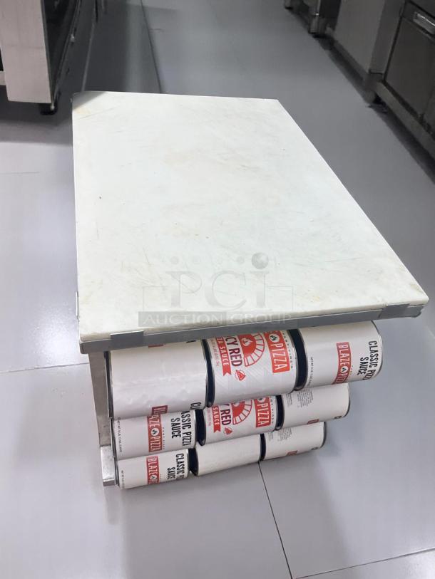 White rectangular cutting board on a stand made of stacked cans labeled "Classic Pizza Sauce." Lightly used condition.