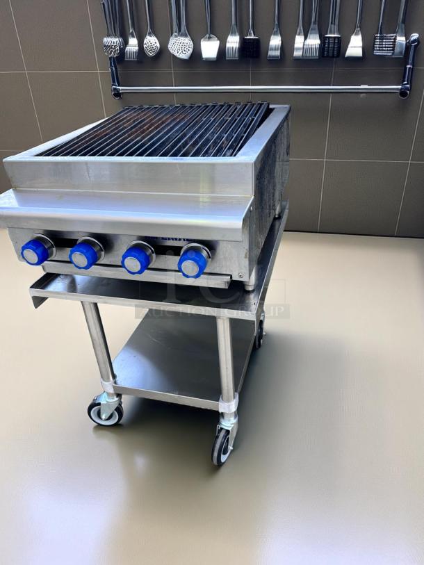 Commercial stainless steel grill with four blue knobs on a wheeled cart. Used condition, visible grill marks. Hanging utensils above.
