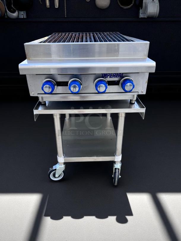 Stainless steel commercial grill with three blue knobs, labeled "Imperial." Mounted on a wheeled stand with a lower shelf.