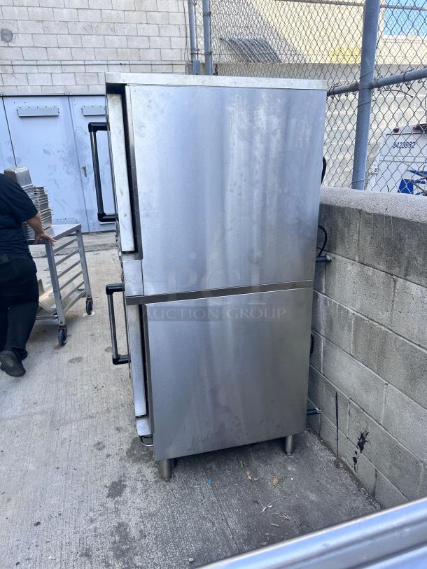 Commercial stainless steel double-door refrigerator, outdoor setting, minor surface wear, sturdy handles, industrial design.