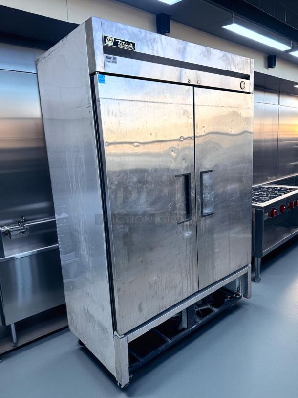 Stainless steel True two-door commercial refrigerator, visible brand label, with signs of wear, in a commercial kitchen setting.