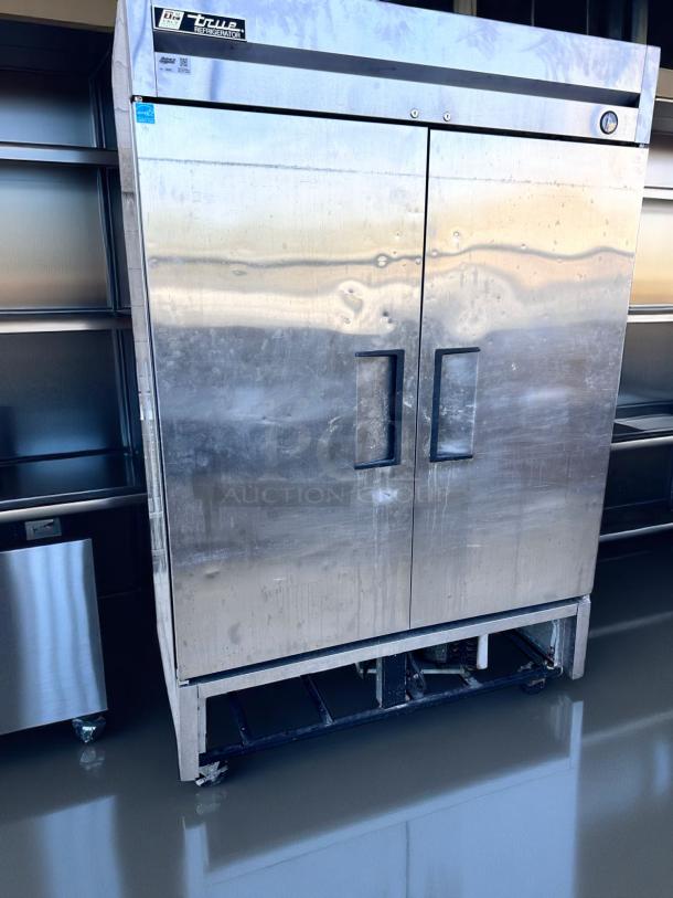 True commercial refrigerator, stainless steel, double doors, visible wear and scratches, model sticker on top-left.