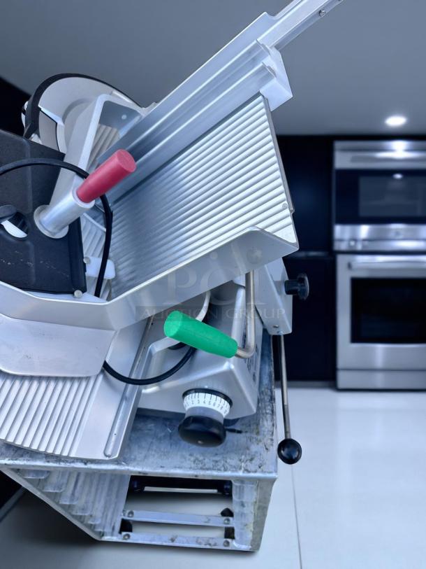Commercial deli meat slicer, stainless steel, with sharp circular blade, red and green handles, and adjustable thickness dial.