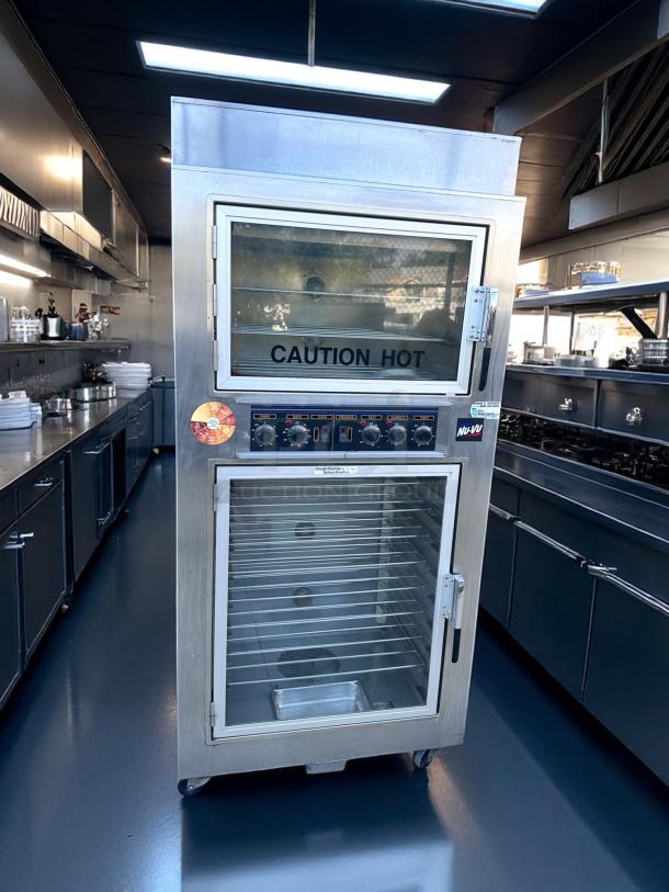 Stainless steel commercial warming oven, Nu-Vu brand, with clear glass doors labeled "Caution Hot," adjustable controls, on wheels.
