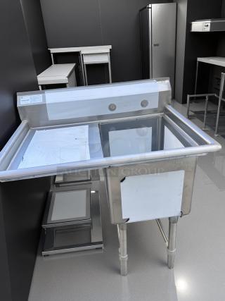 Stainless steel commercial kitchen sink with backsplash, unused condition. Includes shelving and visible quality inspection labels.