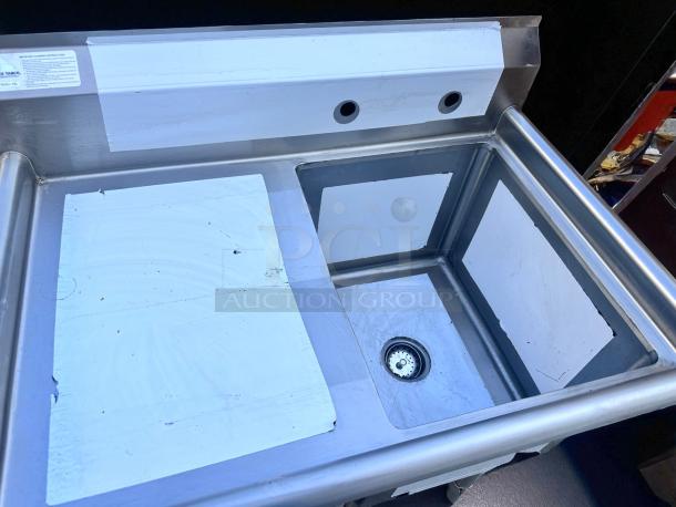 Stainless steel utility sink with dual holes for fixtures, deep basin, and visible cleaning instructions label. Slight wear visible.