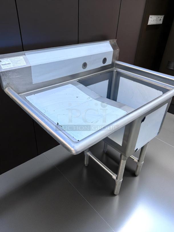 Stainless steel utility sink with backsplash and two faucet holes, supported by sturdy legs. Protective film partially visible.