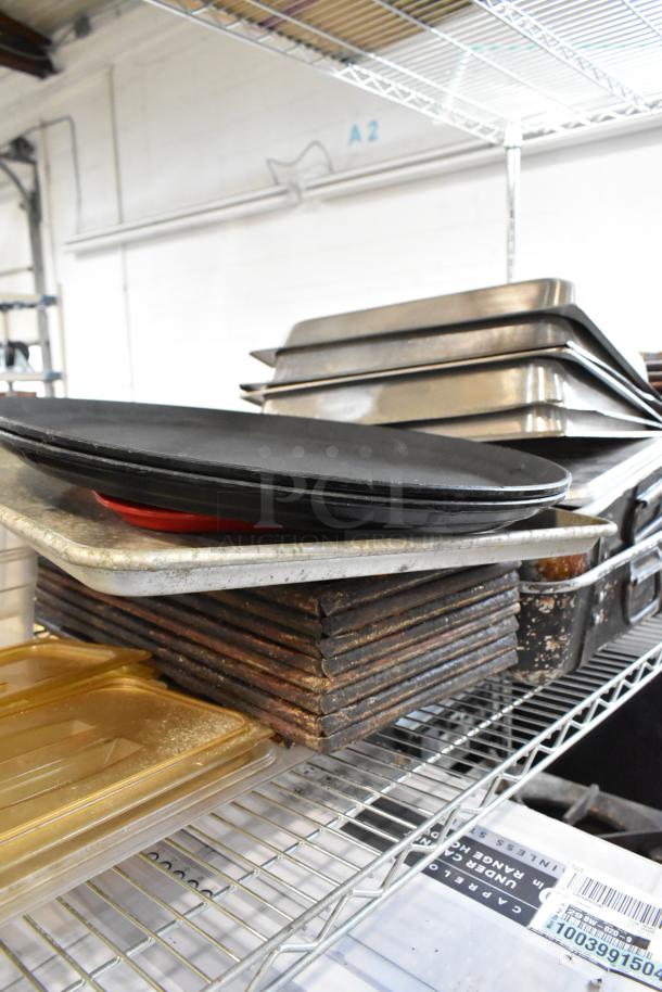 Stacked metal baking trays and pans on wire shelving, various sizes and used condition. Industrial kitchenware.