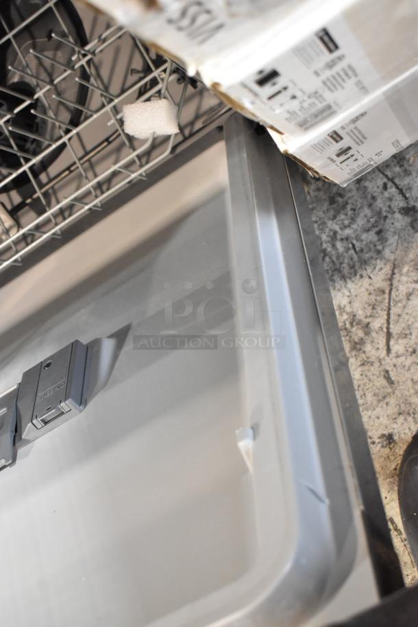 Dishwasher interior with stainless steel tub and rinse aid dispenser, showing packaging materials; appears unused.