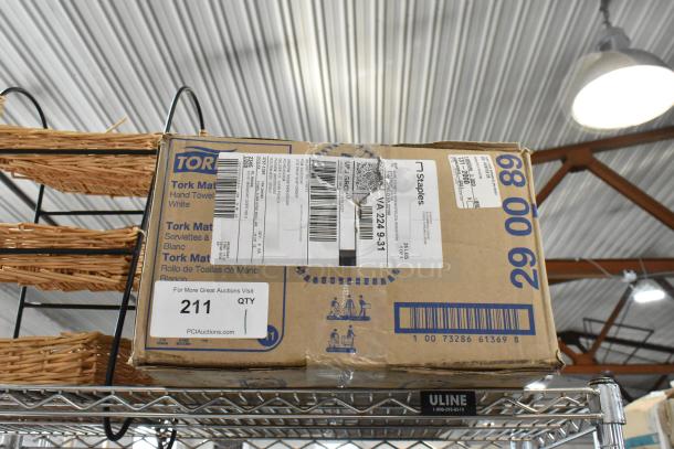 Cardboard box of Tork hand towels, with shipping and auction labels, on a metal shelf. Box in fair condition.