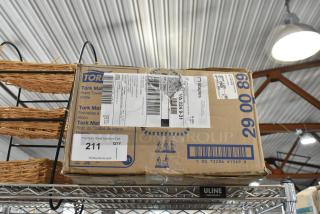 Cardboard box of Tork hand towels, with shipping and auction labels, on a metal shelf. Box in fair condition.