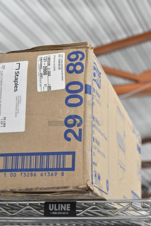 Cardboard box labeled "Tork Matic," weight 25 lbs. Features Staples shipping label. Appears in good condition on metal shelf.