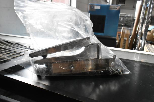Stainless steel component in a sealed plastic bag, includes screws, appears unused, possibly a machine part.