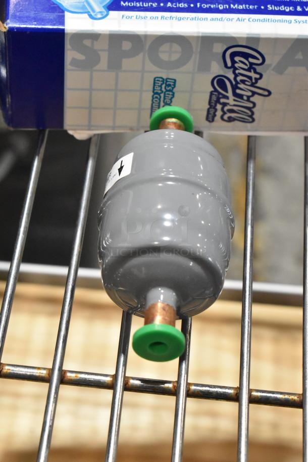 Gray Catch-All filter drier for refrigeration systems, shown new with original packaging. Arrow label indicates flow direction.